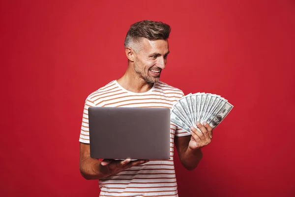 Image of handsome guy in striped t-shirt smiling while holding fan of money cash and laptop isolated over red background