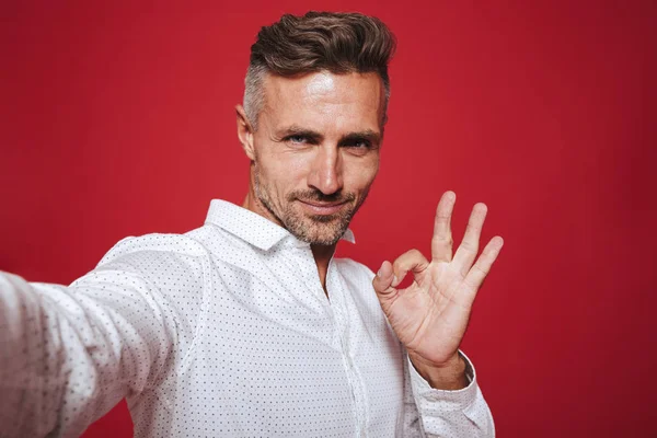 Mutlu esmer adam 30'lu anız selfie fotoğraf çekme ve ok işaret gösteren beyaz gömlek ile kırmızı arka plan üzerinde izole