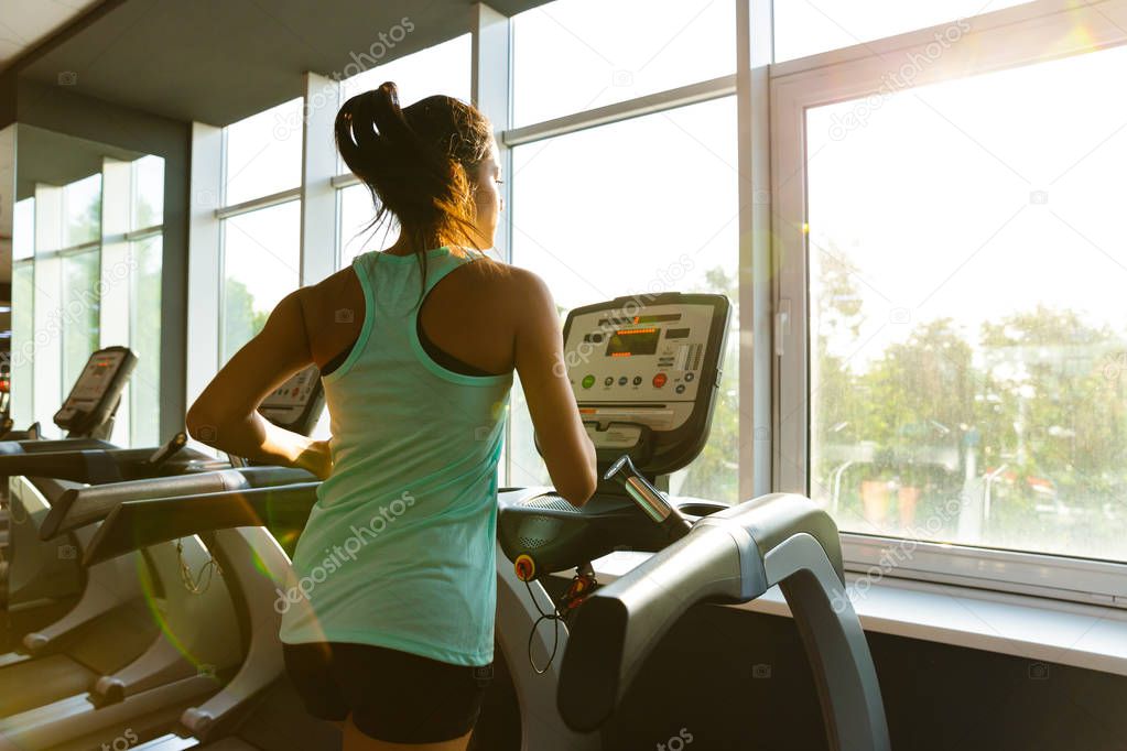 Side view of Young motivated sports woman running on treadmill near the window in gym