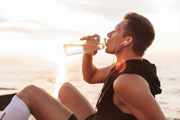 屋外でイヤホン飲料水と音楽を聴くビーチ ハンサムな強い若いスポーツマンの写真 — ストック写真