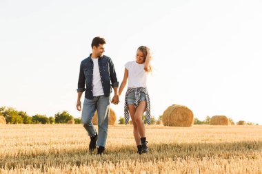 Fotoğraf güzel çift erkek ve kadın haystacks grup ile altın alan aracılığıyla güneşli gün boyunca yürüyüş