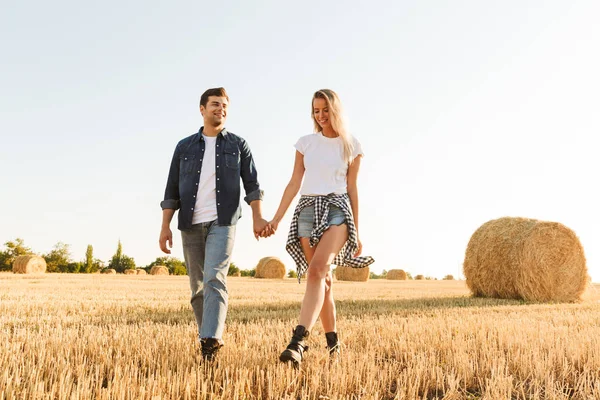 若いカップルの男と晴れた日の間に干し草の束を持つゴールデン フィールドを歩く女性の写真 — ストック写真