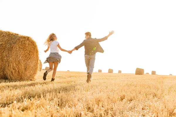 スタイリッシュなカップル男女晴れた日の間に干し草の束を持つゴールデン フィールドを歩きながら楽しんでの写真 — ストック写真