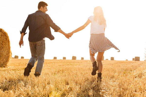 Photo of joyous man and woman running through golden field with bunch of haystacks during sunny day