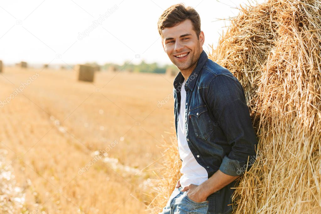 Image of joyful man 30s walking through golden field and standing near big haystack during sunny day