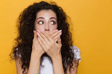 Görüntü gülümseyen ve izole sarı arka plan üzerinde avuç içi ile ağız kapsayan kıvırcık saçlı korkmuş ya da şok kadın 20'li
