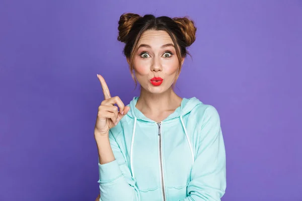 Portrait of funny young girl with two buns pointing finger upward like have idea isolated over violet background in studio