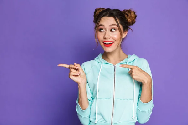 Portrait of caucasian young girl with two buns pointing fingers aside at copyspace isolated over violet background in studio