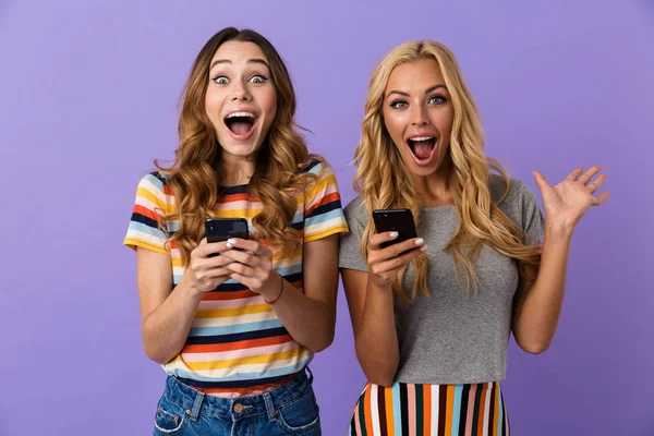 Две Взволнованные Юные Подруги Стоят Изолированные Фиолетовом Фоне Используя Мобильные — стоковое фото