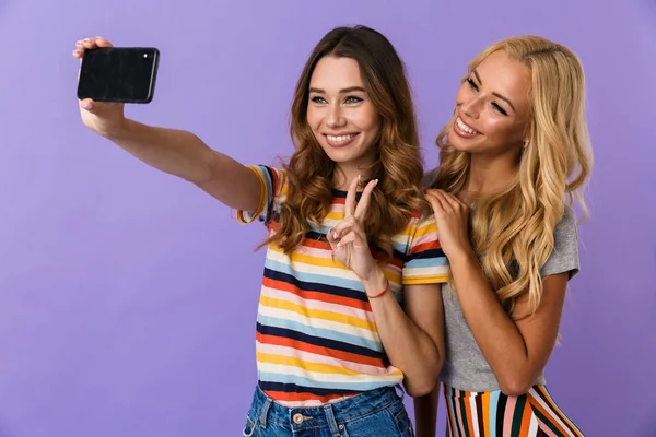 两个漂亮的微笑的年轻女孩的朋友站在紫色背景隔绝 采取自拍与手机 — 图库照片