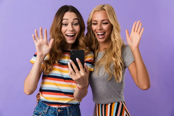 かなり陽気な若い女の子友達を つ紫色の背景に分離立って携帯電話に手を振って — ストック写真
