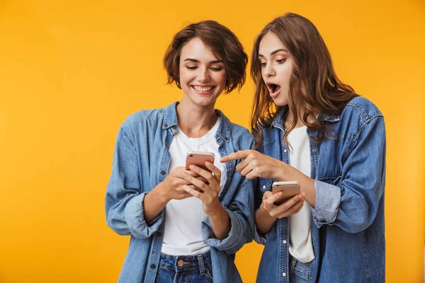 携帯電話を使った黄色の背景に分離されたポーズのショックを感情的な若い女性の友人の写真 — ストック写真