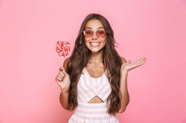 Fotoğraf gülüyor ve pembe arka plan üzerinde izole lolipop tutan Avrupa kadın 20'li yaşlarda giyen güneş gözlüğü