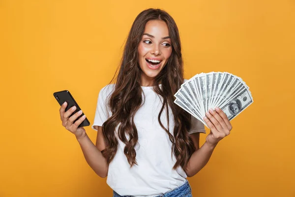 一个高兴的年轻女孩的肖像站在黄色背景 持有钞票 使用手机的长黑发头发 — 图库照片
