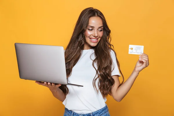 黄色の背景の上に立って ラップトップ コンピューターを保持している プラスチック製のクレジット カードを示す長いブルネットの髪で陽気な若い女の子の肖像画 — ストック写真