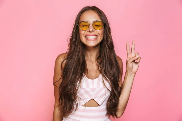 ブルネットの女性 代身に着けているサングラスと笑ってピースサイン ピンク背景に分離を示す写真 — ストック写真