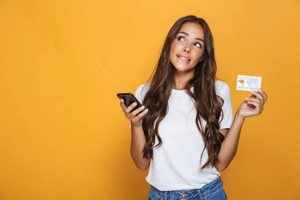 一个沉思的年轻女孩与长黑发头发站立在黄色背景 手持手机 显示塑料信用卡的肖像 — 图库照片#