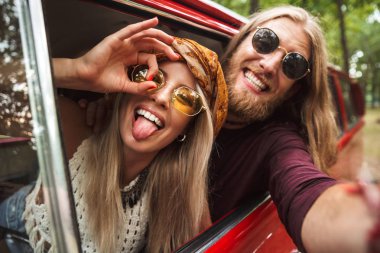 Photo of caucasian hippie couple smiling and fooling around while driving retro minivan in forest