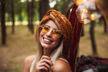 Şık aksesuarlar orman kampında istirahat ederken gülümseyen giymiş sarışın gezgin kadın fotoğrafı