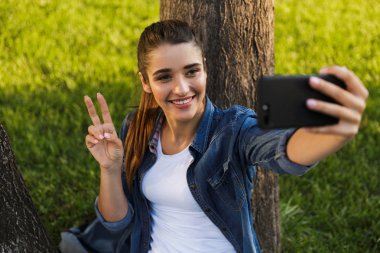 İnanılmaz güzel ve genç birbayan öğrenci parkta görüntüsünü almak bir selfie tarafından cep telefonu barış hareketi ile.