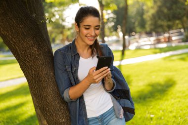 Görüntü neşeli genç güzel kadın öğrencinin cep telefonuyla bir kenara park seyir içinde yürüyüş.