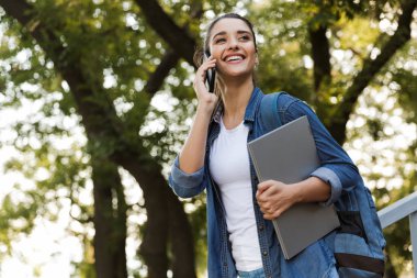 Cep telefonuyla konuşurken dizüstü bilgisayar açık havada tutan duran genç ve güzel beyaz kadın görüntüsünü.
