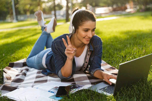 惊人的快乐的女人学生的形象躺在户外在公园使用笔记本电脑听音乐做和平手势 — 图库照片