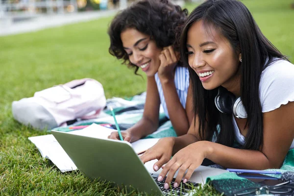 Two cheerful young girls students laying on a grass at the campus, studying with laptop computer and textbooks