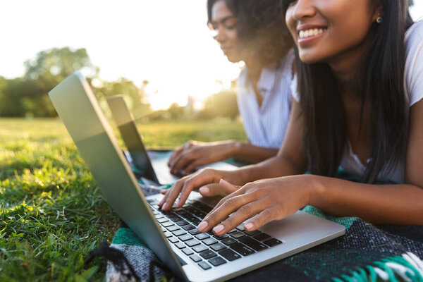Крупный план двух улыбающихся юных девушек, лежащих на траве в кампусе, занимающихся с ноутбуками
