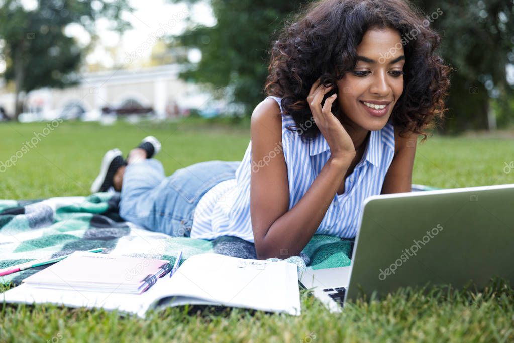 Happy african girl student laying on a grass at the campus, studying, using laptop