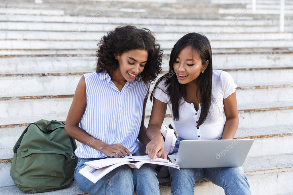 Two cheerful young girls students sitting on steps at the campus, studying with laptop computer and textbooks