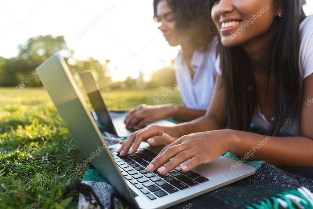 Close up of two smiling young girls students laying on a grass at the campus, studying with laptop computers