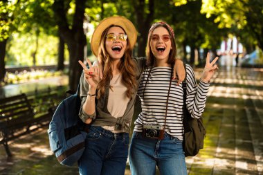 Heyecanlı güzel genç mutlu kadın arkadaşlar açık havada yürümek ile görüntü gösteren barış jest sırt çantaları.