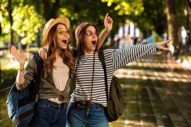 Heyecanlı genç ve güzel mutlu kadın arkadaşlar açık havada yürümek ile görüntü işaret sarılma sırt çantaları.