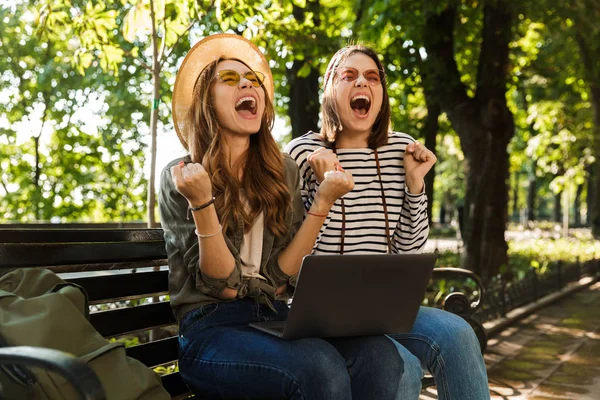 Photo of young happy excited happy ladies friends outdoors sitting using laptop computer make winner gesture.