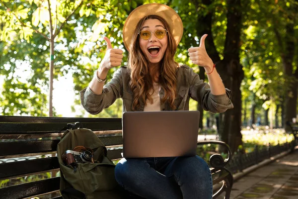 照片的年轻美丽兴奋愉快的妇女户外坐在使用膝上型计算机做竖起大拇指 — 图库照片