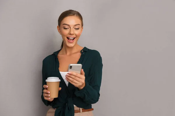 Image of excited beautiful business woman posing isolated over grey wall background drinking coffee using mobile phone.