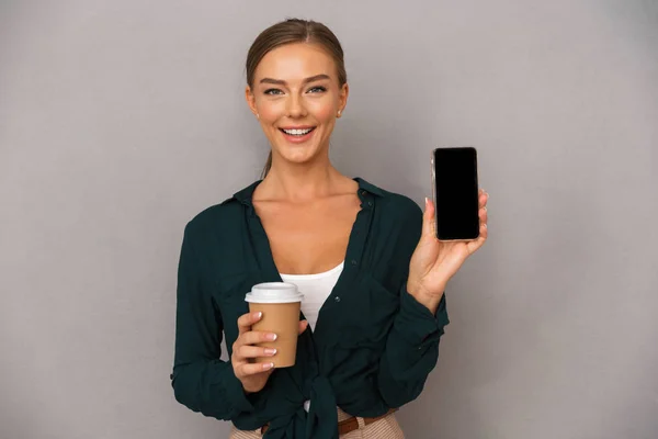 Image of excited beautiful business woman posing isolated over grey wall background drinking coffee showing display of mobile phone.