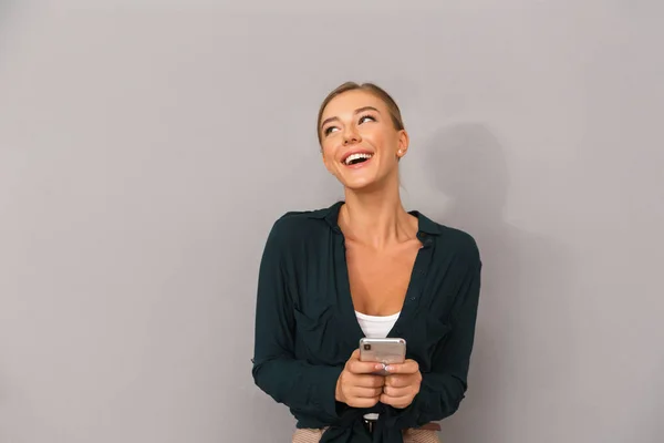 携帯電話を使用して灰色の壁背景に分離された陽気なハッピー ビジネス若い女性がポーズの画像 — ストック写真