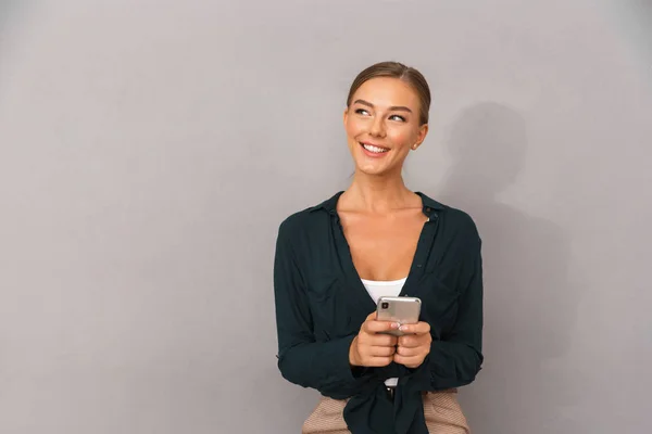 携帯電話を使用して灰色の壁背景に分離された陽気なハッピー ビジネス若い女性がポーズの画像 — ストック写真