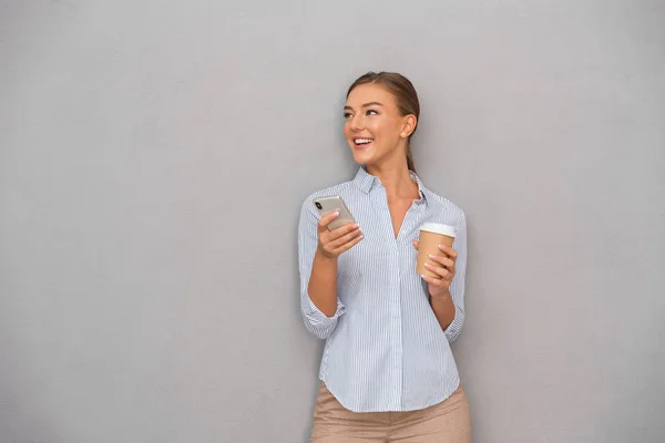 コーヒーを飲みながら携帯電話を使用して灰色の壁背景に分離された幸せなビジネス若い女性がポーズの画像 — ストック写真