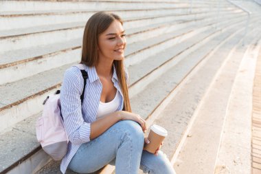 Gülümseyen bir genç kız açık havada merdivenlerde otururken paket servisi olan restoran kahve tutan sırt çantası ile yakın