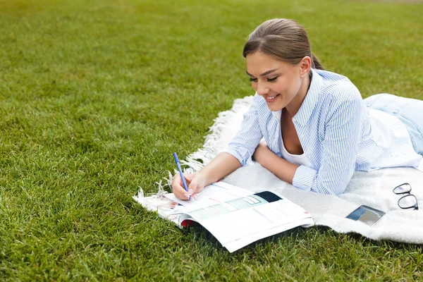 Cheerful young girl spending time at the park, studying, laying on a blanket on grass