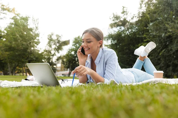 微笑的年轻女孩花时间在公园 铺设在毯子与笔记本电脑 在手机上交谈 — 图库照片