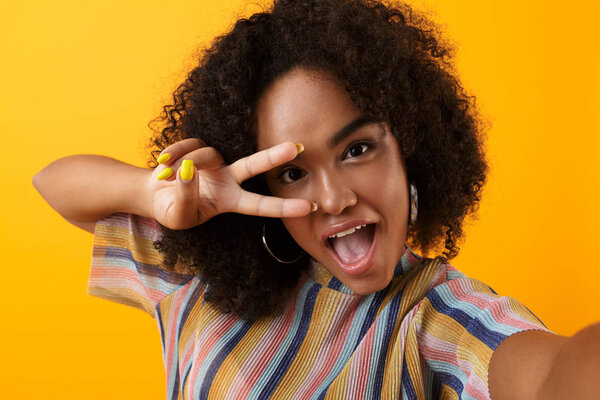 Image of beautiful young happy african cute girl posing isolated over yellow background take a selfie with peace gesture.