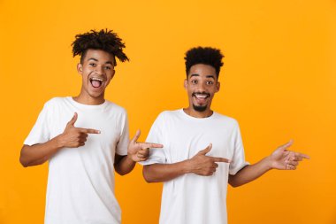 İki mutlu erkek Afrika arkadaşlar sarı arka plan üzerinde izole ayakta t-shirt uzağa işaret içinde