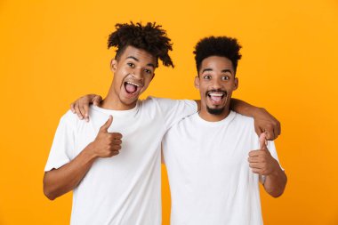 İki neşeli erkek Afrika arkadaşlar başparmak gösterilen sarı arka plan üzerinde izole ayakta t-shirt