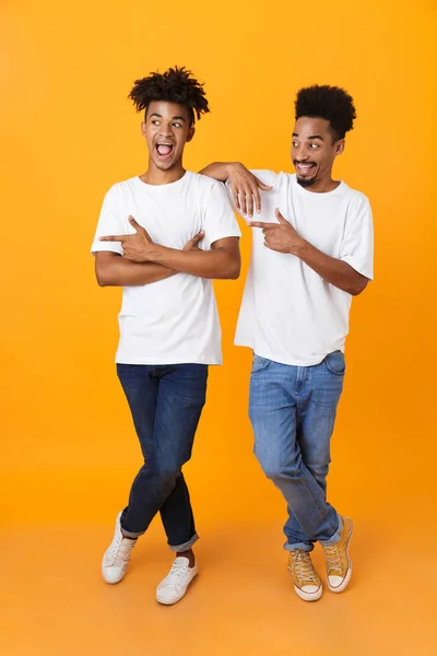 Full length two excited male african friends in t-shirts standing isolated over yellow background, pointing away