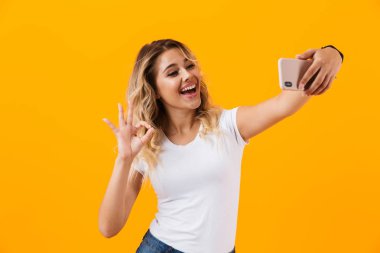 Görüntü gülümseyen ve sarı arka plan üzerinde izole Smartphone cep telefonu ile selfie fotoğrafta şirin sarışın kadın