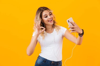 Görüntü gülümseyen ve cep telefonu sarı arka plan izole selfie fotoğrafta kulaklık giyen pozitif kadın